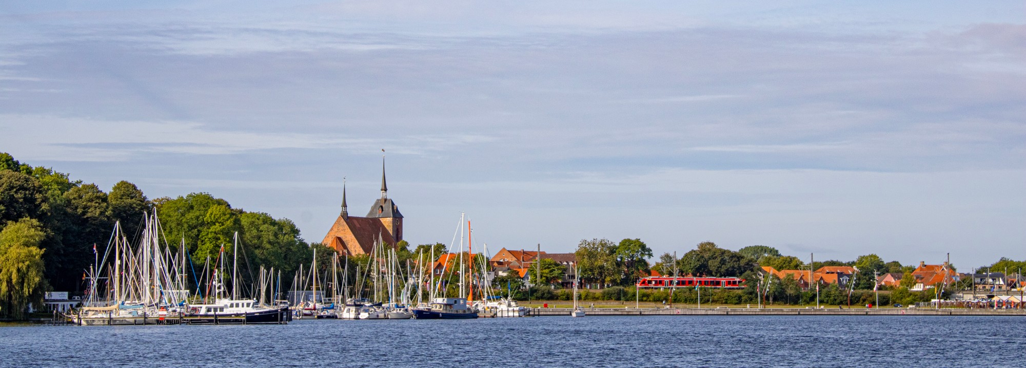 Panorama vom Obereiderhafen Rendsburg an einem sonnigen Tag: ein Yachthafen und eine belebte Promenade im Vordergrund, hinter Bahngleisen die Gebäude der Altstadt mit einer Kirche